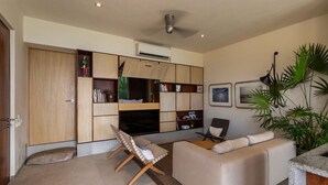 Una sala moderna con un sofá, una mesita y un banco. Se ve una estantería con diversos objetos y un ventilador de techo.