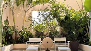 Un patio con bancos blancos y una mesa redonda central, rodeado de abundante vegetación y una pared color beige.