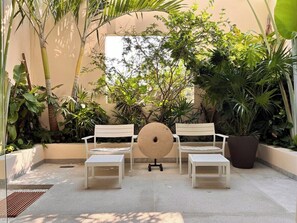 A patio with white benches and a central round table, surrounded by lush greenery and a beige wall.