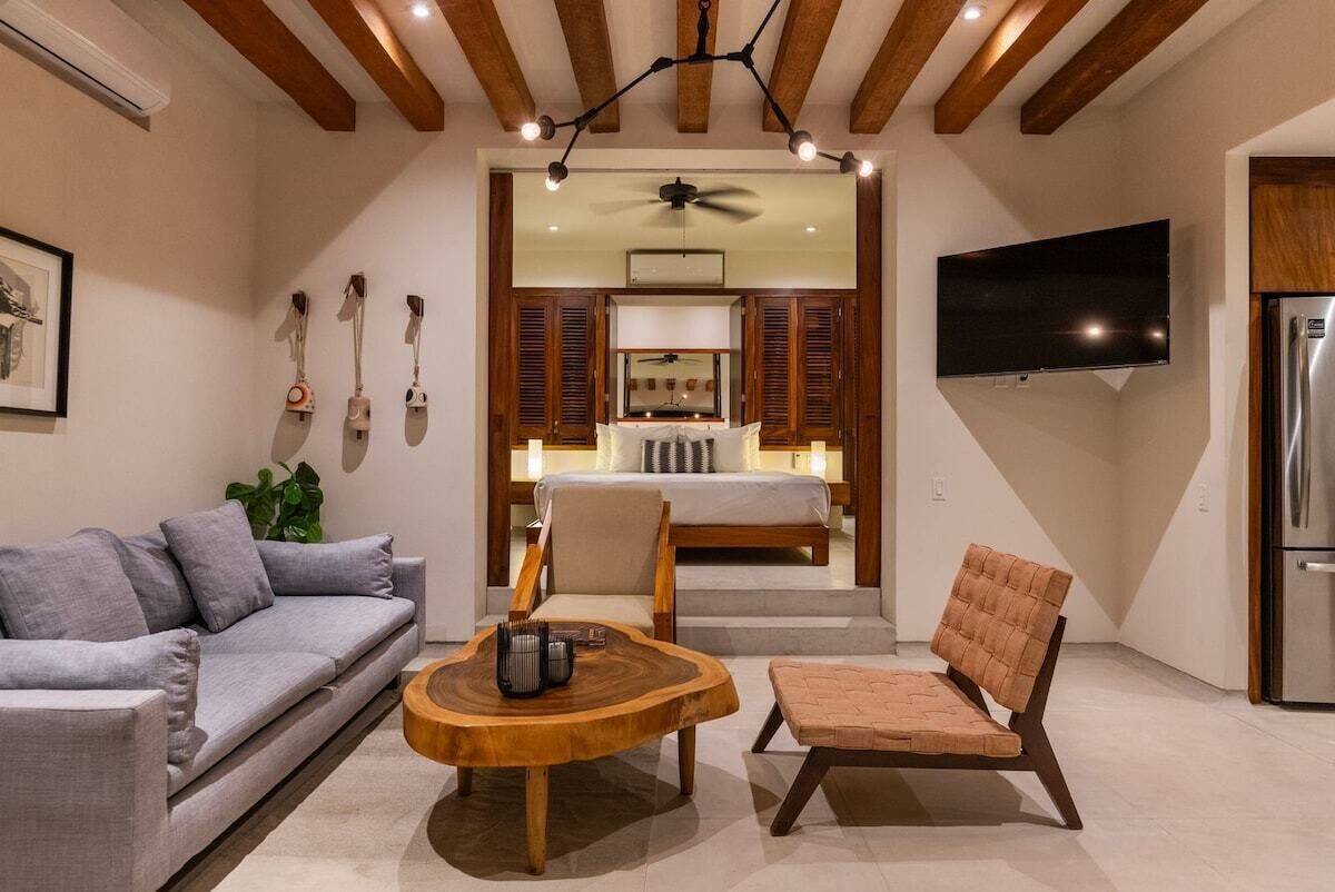 A modern living room with a sofa, a wooden coffee table, and a cushioned chair. A television is mounted on the wall, and a ceiling fan is visible.