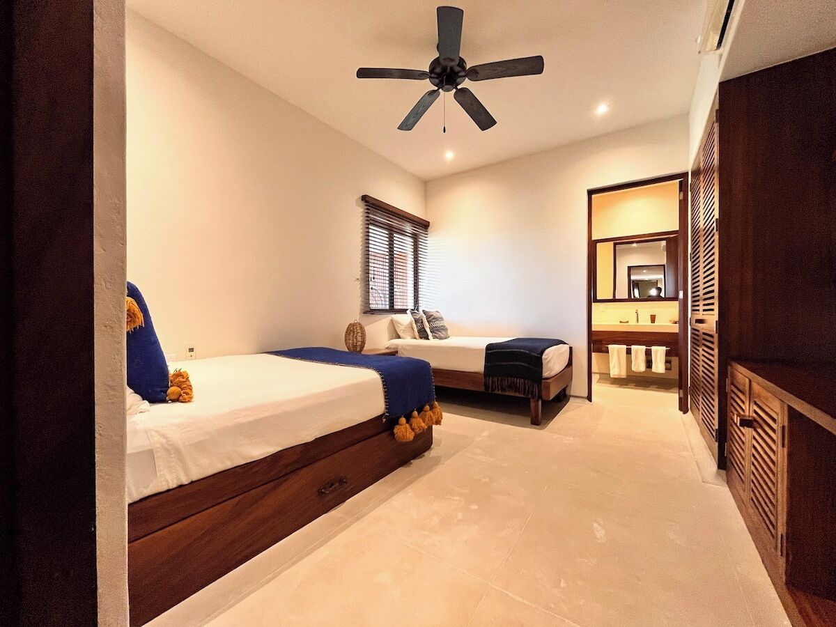 A bedroom with two beds, a ceiling fan, a bathroom with a mirror, and a wooden wardrobe.