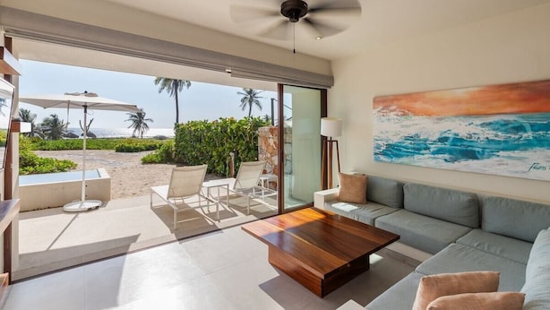 A modern living room with a sofa, coffee table, and chairs, a ceiling fan, and a painting of a beach scene. - Beachfront 3BR Villa I Private Pool + Sand Access (Zihuatanejo)
