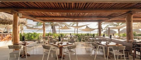 An outdoor dining area with tables and chairs, a wooden pergola overhead, and a view of the beach and palm trees.