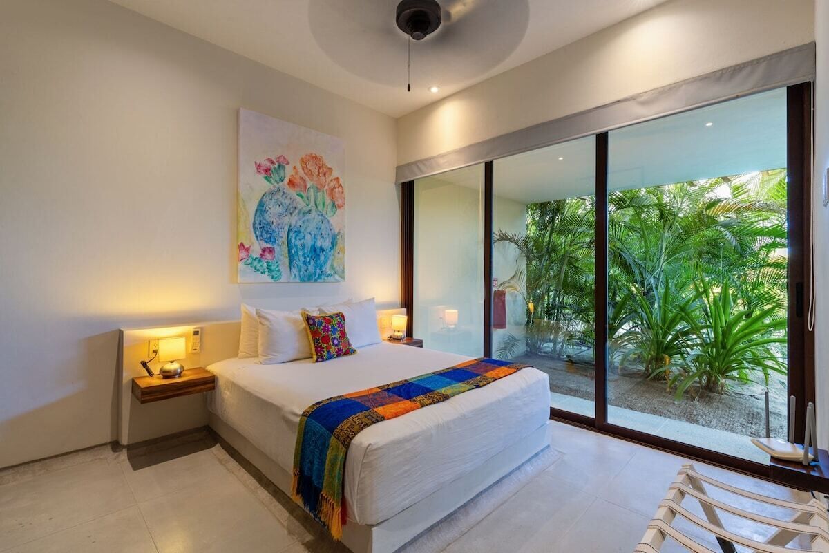 Une chambre d’hôtel moderne avec un grand lit, des lampes de chevet, une œuvre d’art colorée au mur et une vue sur la verdure grâce à de grandes fenêtres du sol au plafond.