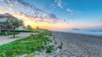 Uma praia com céu claro, areia dourada e um edifício com telhado inclinado.