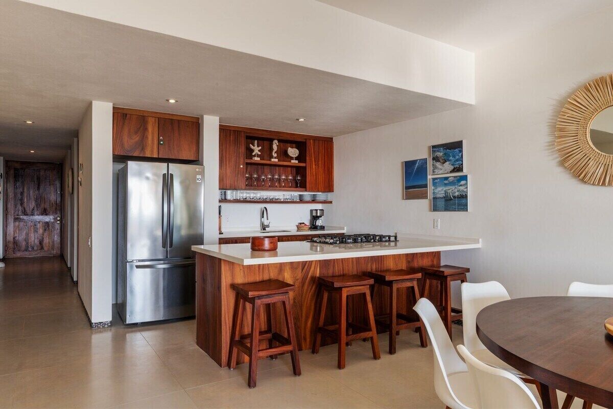 A modern kitchen with a stainless steel refrigerator, wooden cabinetry, a central island with stools, and a round dining table.
