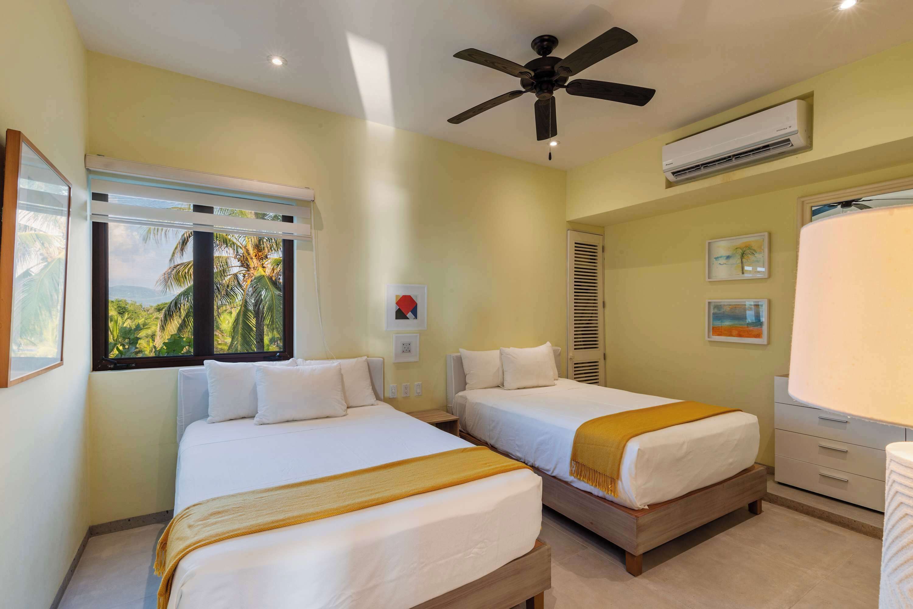 Habitación de hotel con dos camas, ventilador de techo, unidad de aire acondicionado y ventana con vista a palmeras.