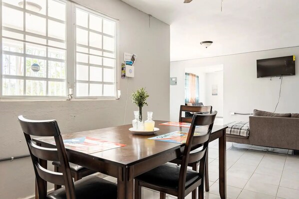 A dining area with a wooden table, four chairs, a vase with flowers, and a plate with a drink. There is a TV mounted on the wall and a sofa in the background.