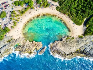 Vista aérea de una playa con aguas turquesas cristalinas, acantilados rocosos y una zona de arena con coches estacionados y sombrillas.