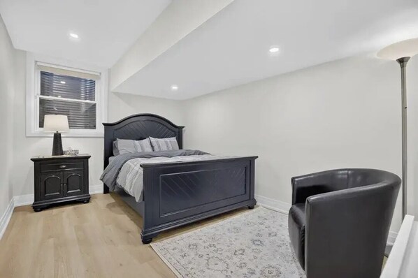 Une chambre à coucher comprenant un lit, une table de chevet, une chaise, une lampe sur pied et une fenêtre avec des stores.