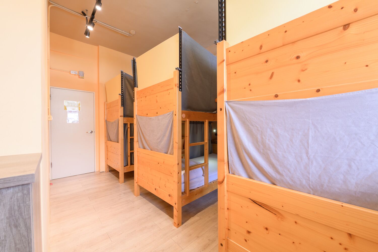 A row of wooden bunk beds with grey sheets, a door with a notice, and a light fixture on the ceiling.