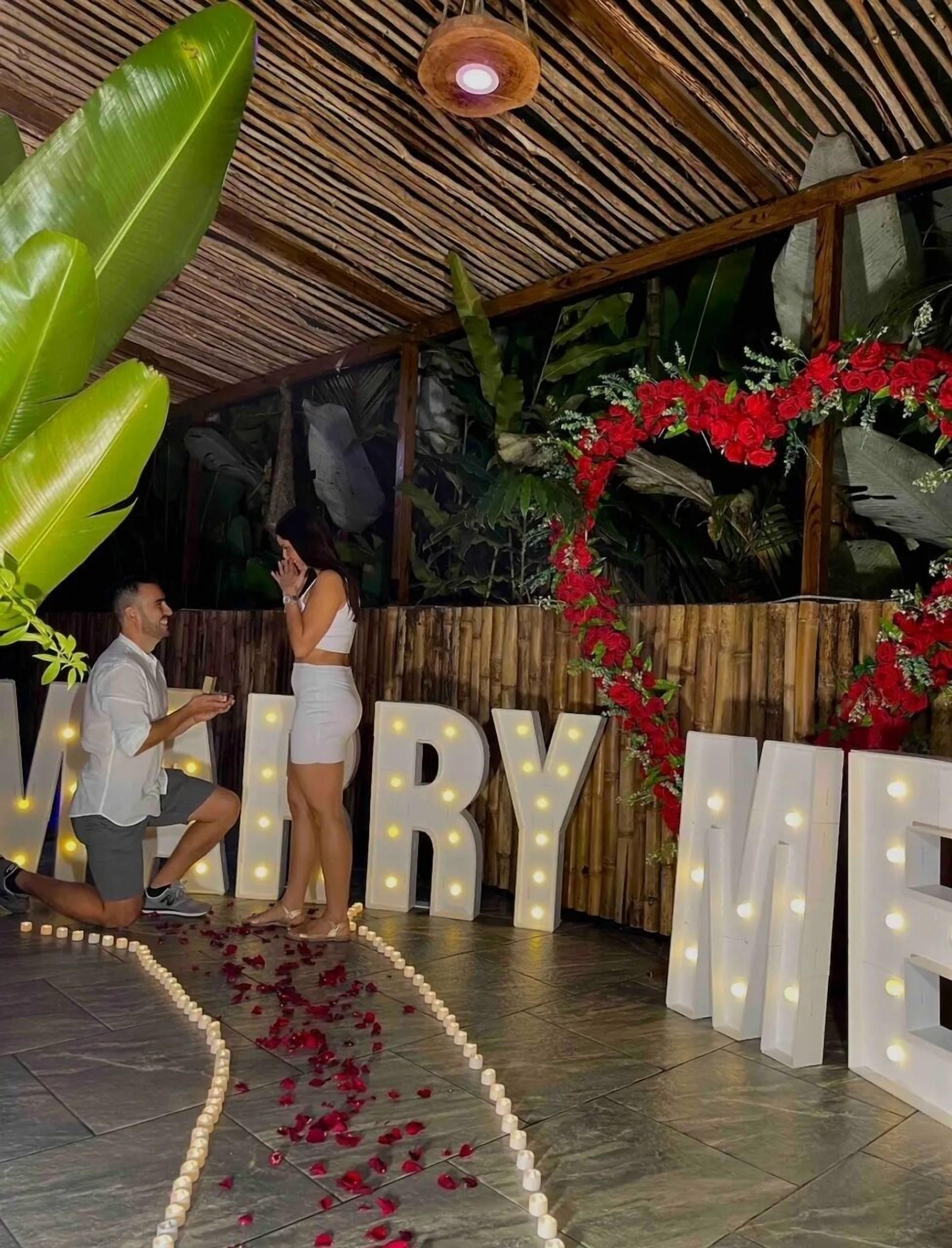Un hombre proponiendo matrimonio a una mujer en un entorno tropical, con un letrero que dice 'MARRY ME', rodeado de follaje e iluminación decorativa.