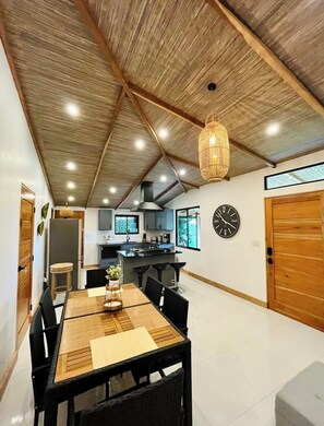 A spacious dining area with a wooden table and chairs, a kitchen in the background, and a large clock on the wall. - Arenal Palms (Peñas Blancas)