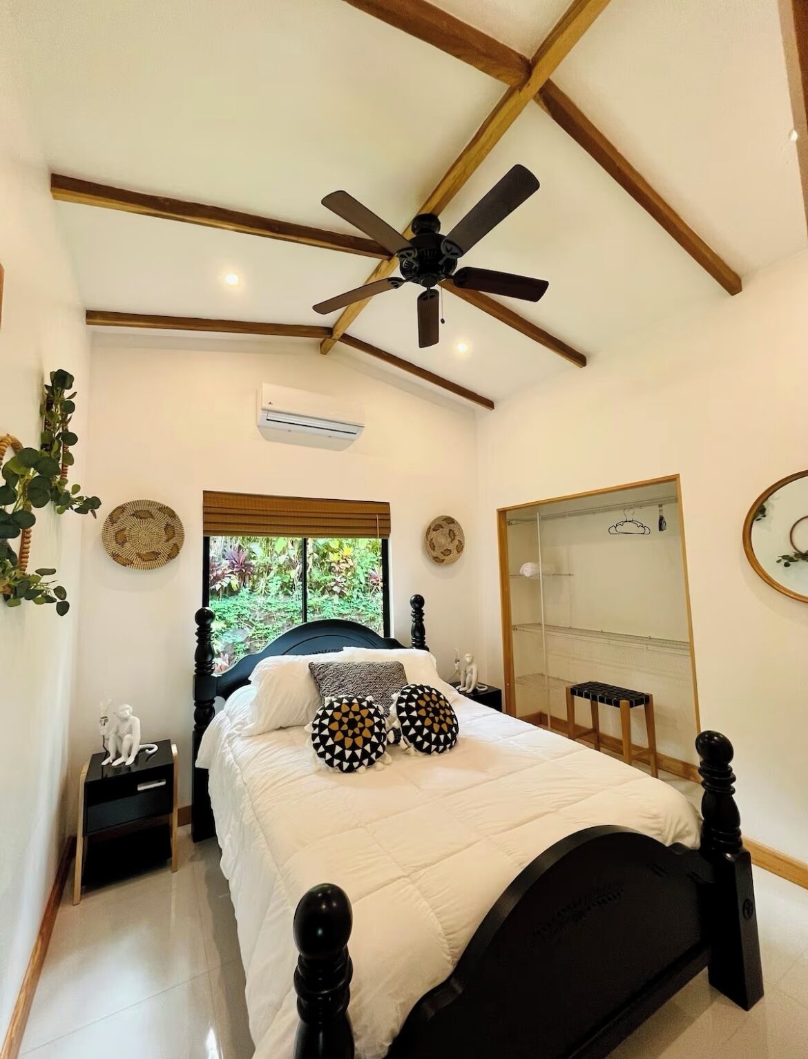 Ein Schlafzimmer mit Bett, Deckenventilator, Fenster mit Blick auf Grünflächen, Nachttisch mit Lampe und Spiegel an der Wand.