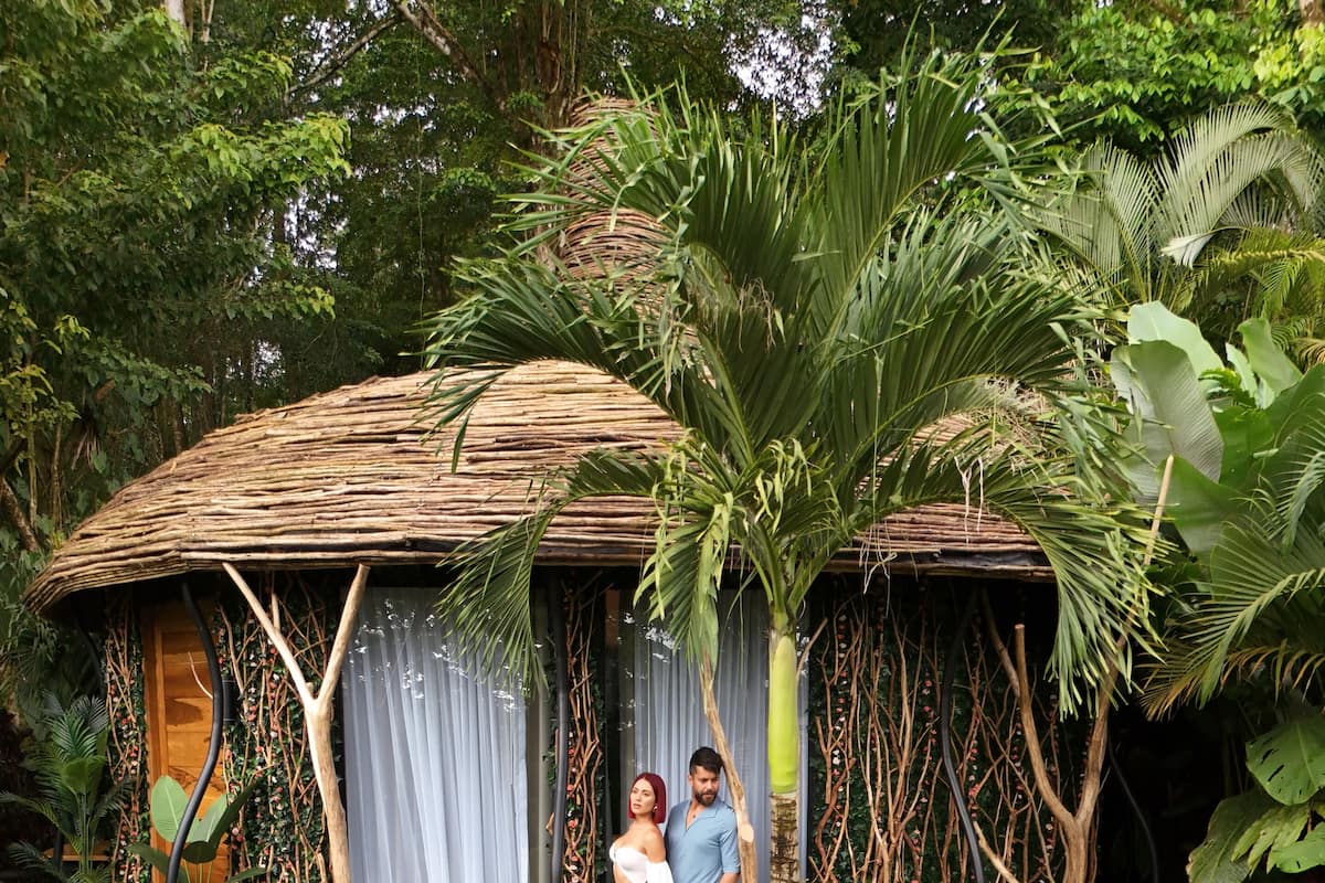 A couple standing on a wooden deck with a thatched roof structure surrounded by lush greenery.