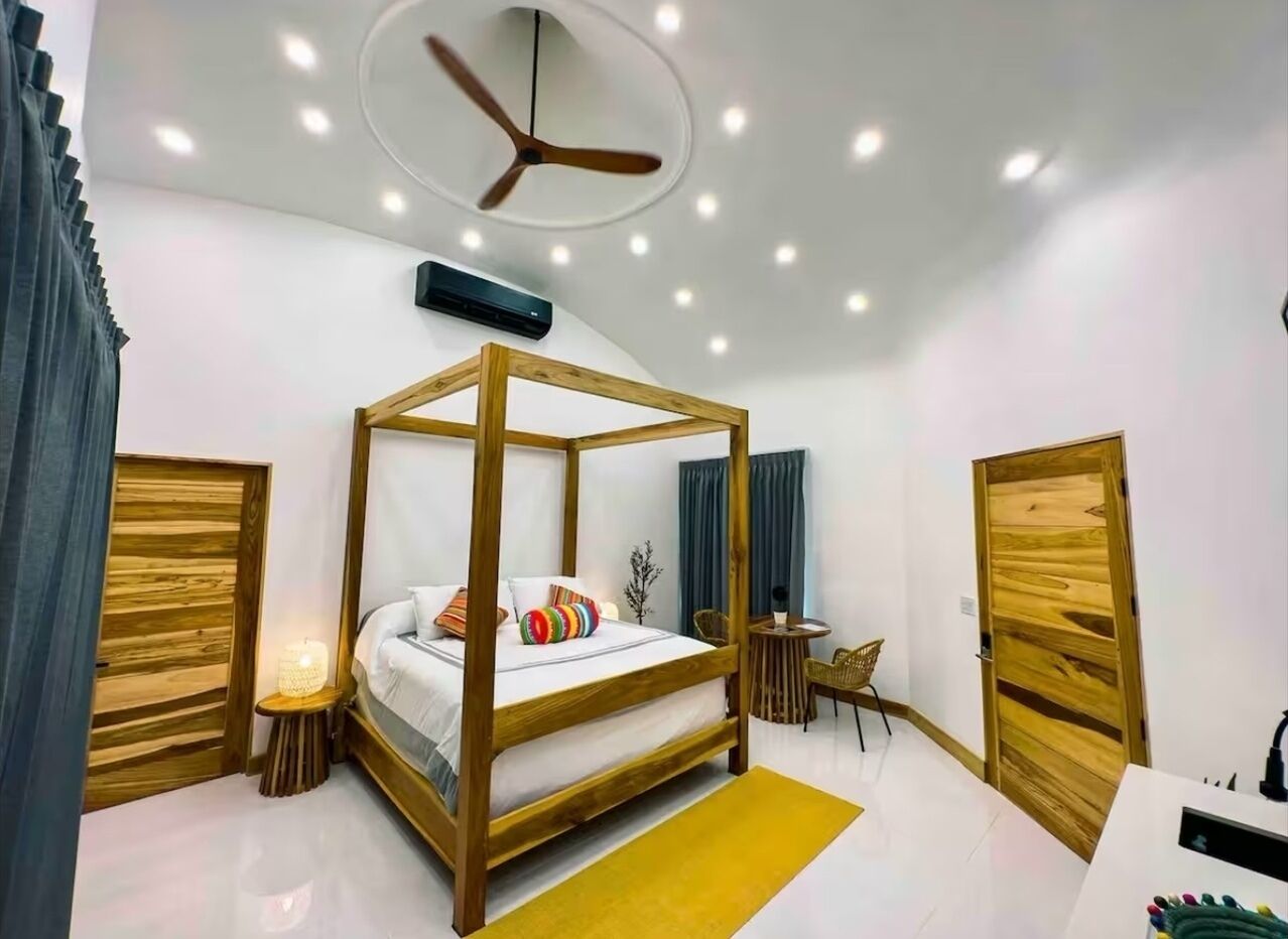 A modern bedroom with a four-poster bed, a ceiling fan, and a small table with a chair.