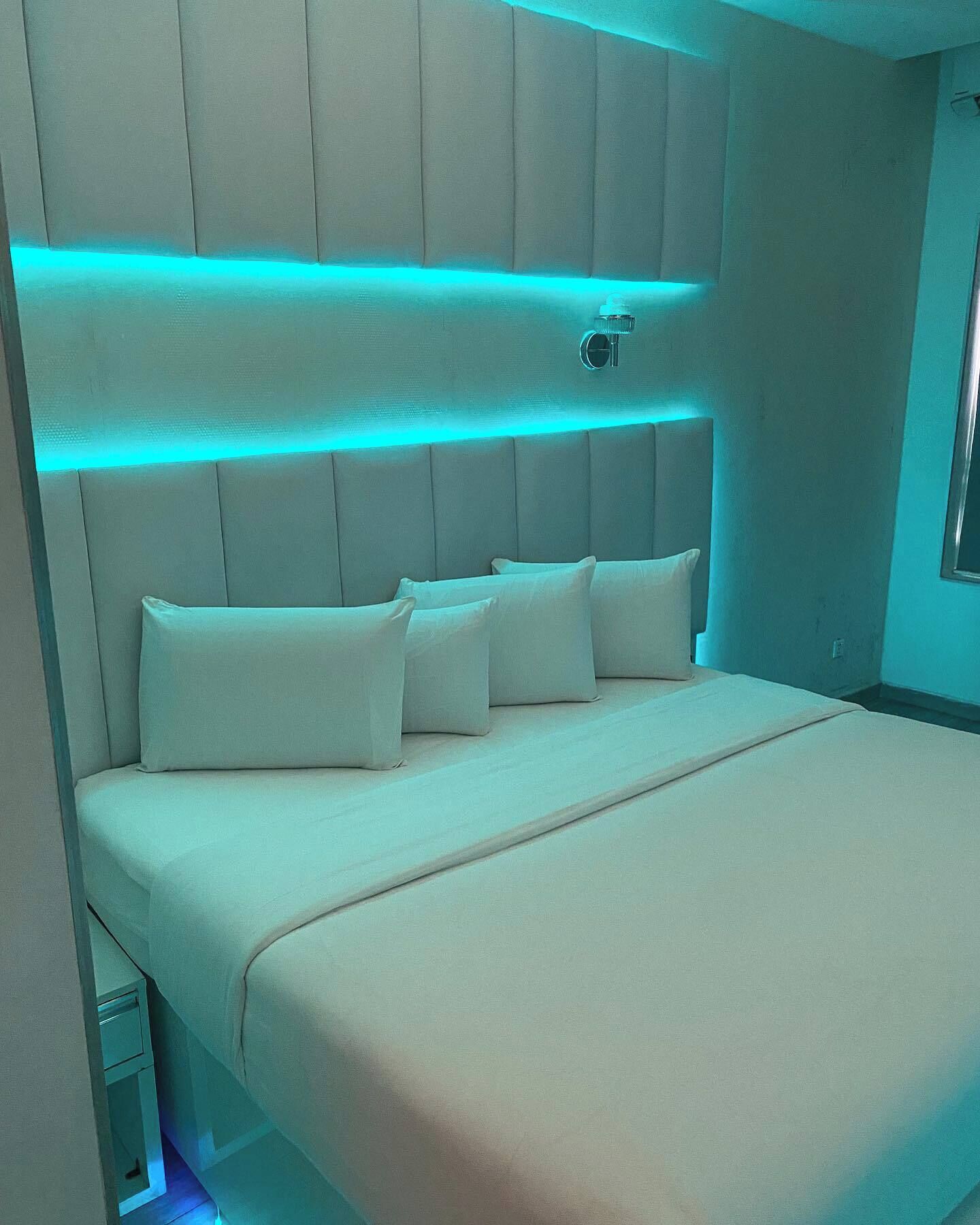 Un dormitorio moderno con cabecera acolchada, cama con ropa de cama blanca y una tira de luz ambiental azul sobre la cama.