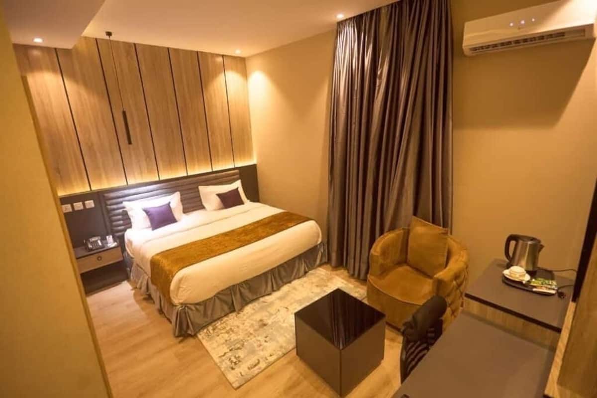 A modern hotel room with a large bed, a leather armchair, a small table, and an air conditioning unit.
