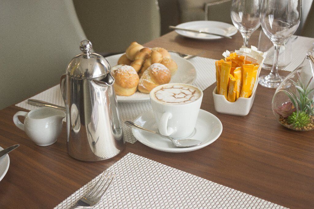 A breakfast setup with a coffee pot, pastries, a cup of coffee, and a small plant on a wooden table.