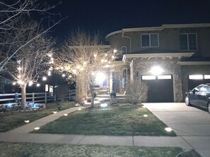 Una propiedad residencial de noche con fachada de piedra, un garaje y un jardín delantero bien iluminado con árboles y luces.