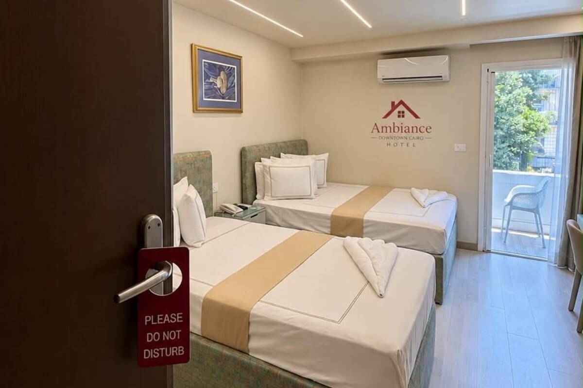 A hotel room with two beds, a door with a 'Please Do Not Disturb' sign, a wall-mounted air conditioner, and a view of the outdoors.