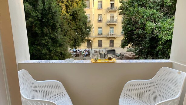 A balcony with two white perforated chairs, a small table with glasses and a tray, and a view of a building with balconies and trees. - Ambiance Downtown Hotel (Cairo)