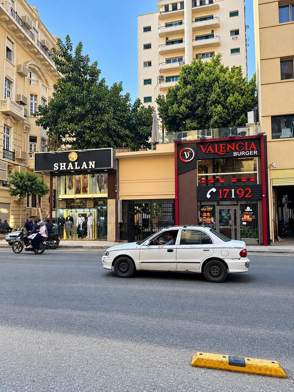 A street view with a white car, a building with a 'Valencia Burger' sign, and a 'Shalan' store.