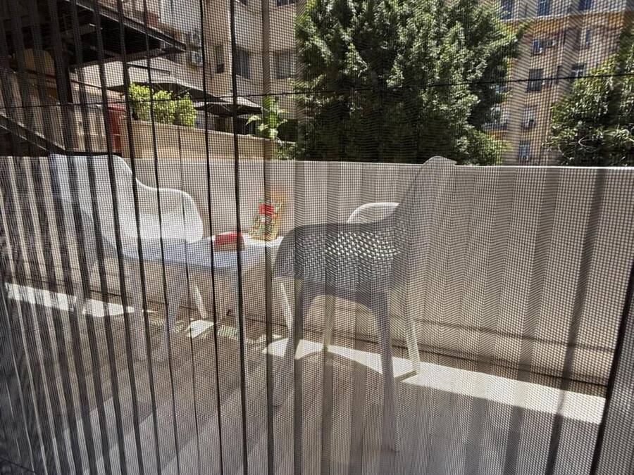 Vista a través de una ventana con mosquitera metálica, mostrando una calle con coches aparcados y edificios al fondo.