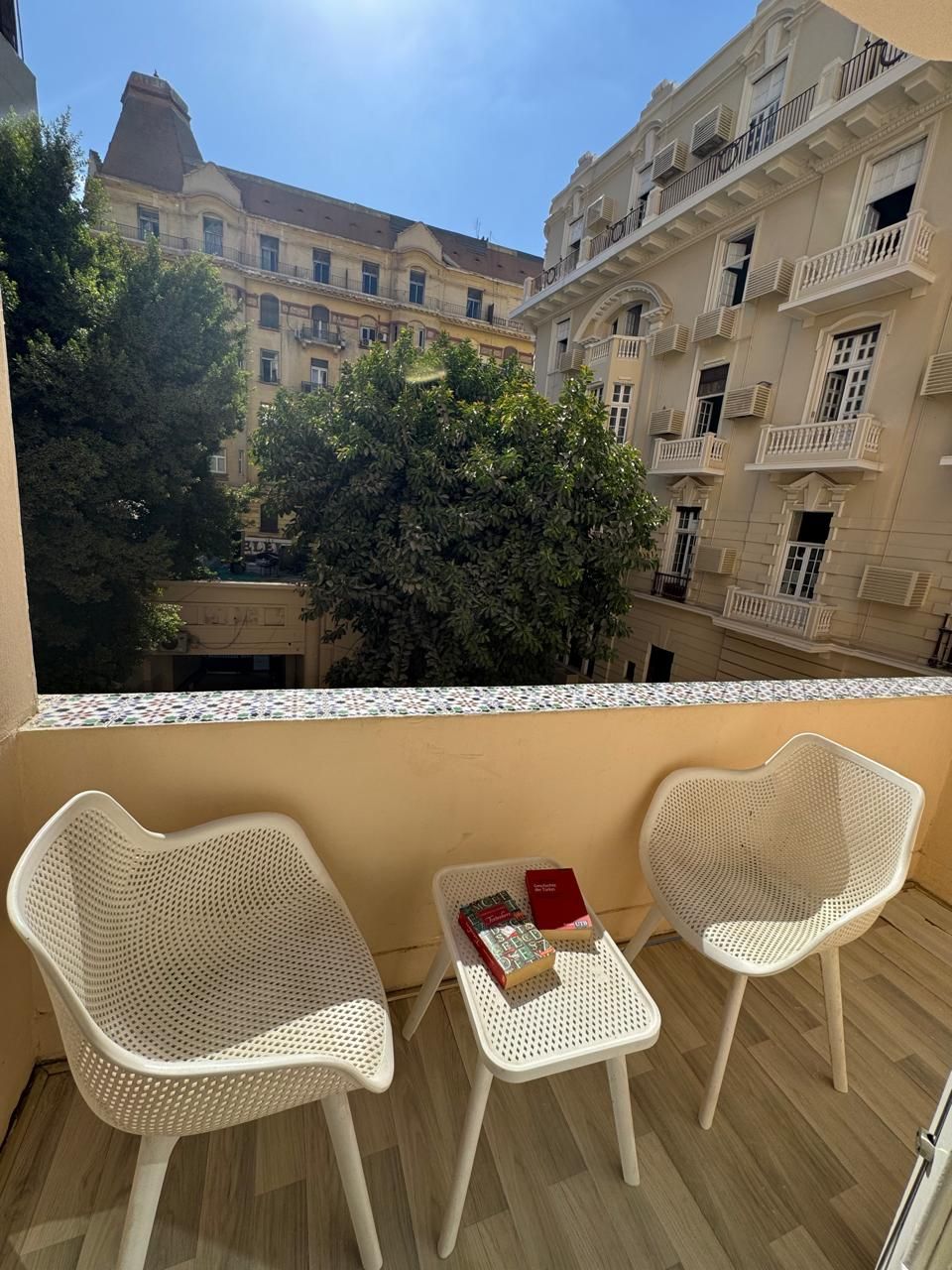 Un balcón con dos sillas de mimbre blancas y una mesita, con vista a un patio con edificios y árboles.