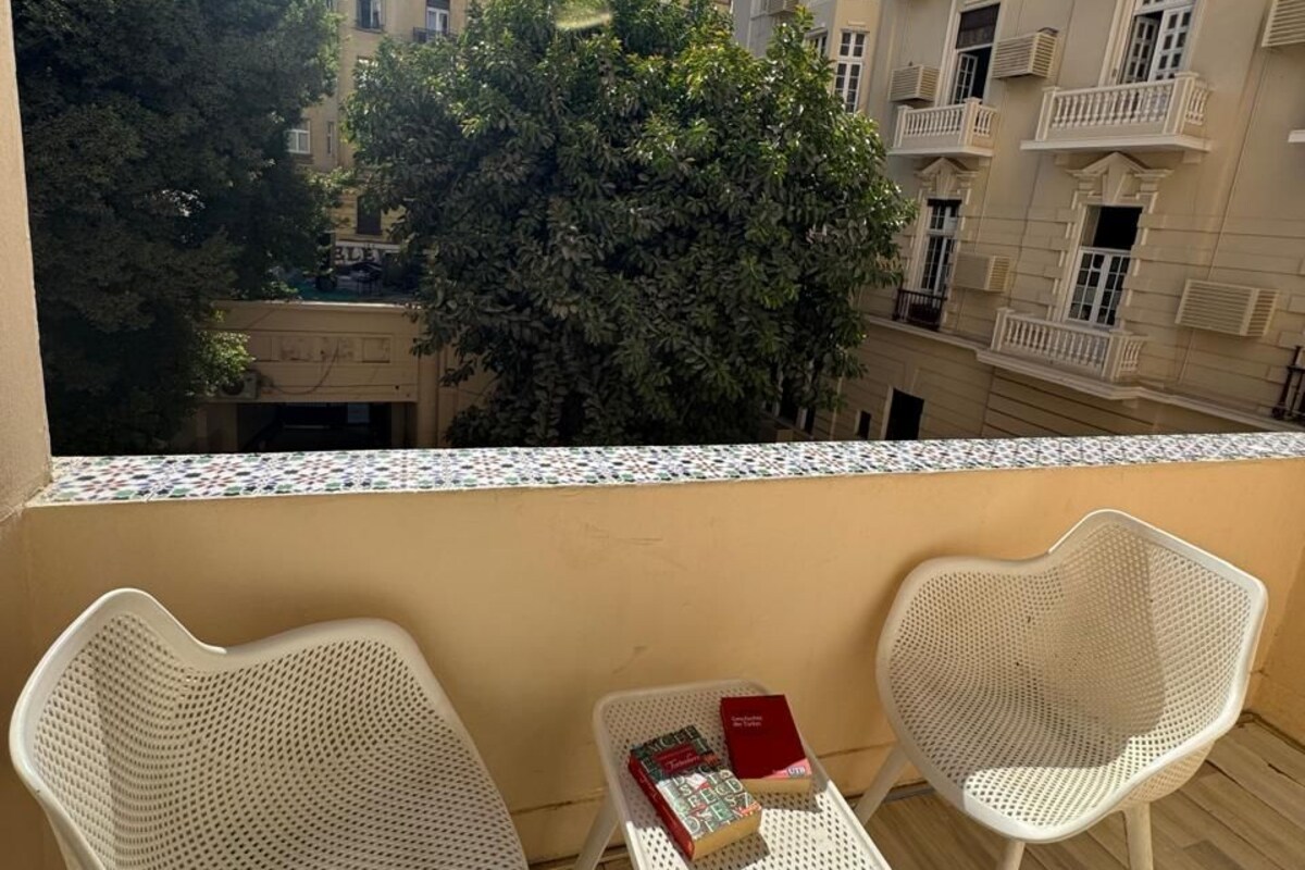 A balcony with two white wicker chairs and a small table, overlooking a courtyard with buildings and trees.