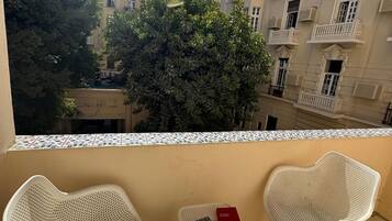 Un balcón con dos sillas de mimbre blancas y una mesita, con vista a un patio con edificios y árboles.
