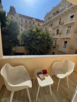 Un balcon avec deux fauteuils en osier blancs et une petite table, donnant sur une cour intérieure bordée d’immeubles et d’arbres.
