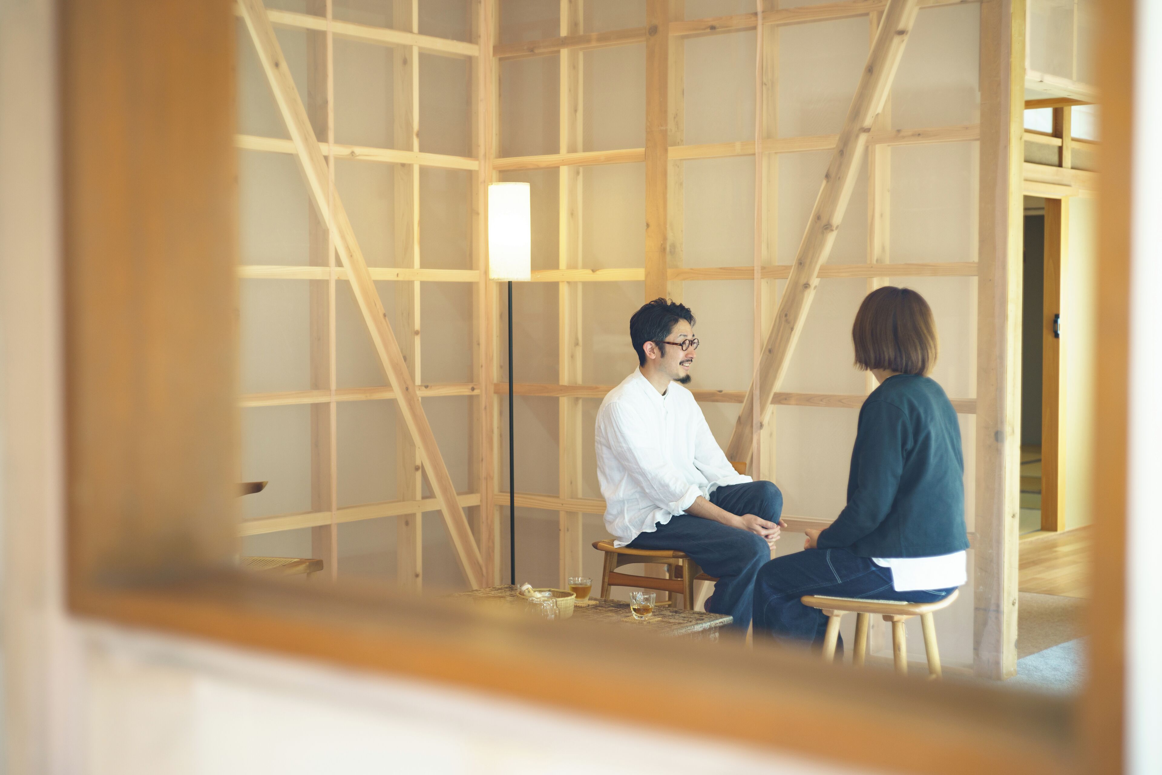 A room with a wooden grid structure, two people sitting on stools, and a small table with a cup and saucer.