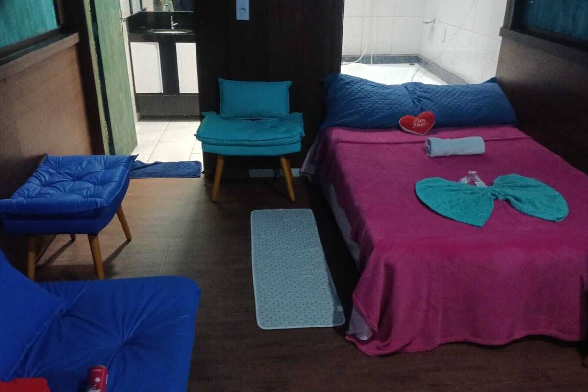 A room with a bed, two blue chairs, a small table, and a view of a kitchen area.