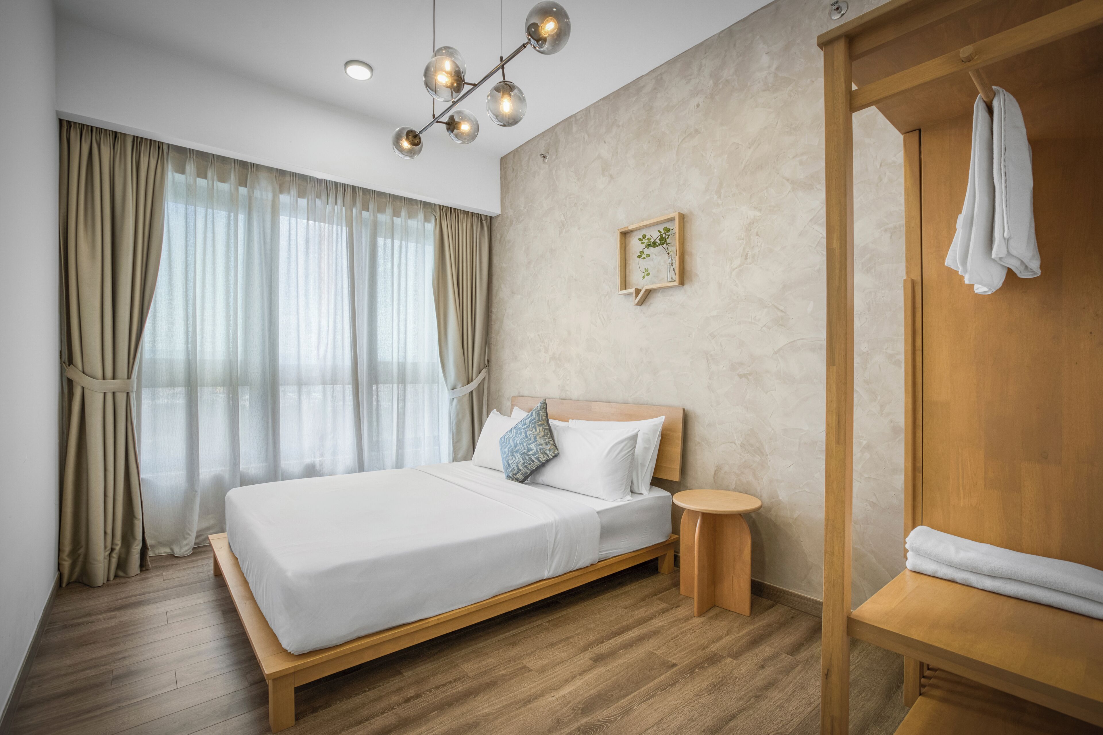 A modern hotel room with a wooden bed, a round wooden side table, a wooden bench, a wooden wardrobe, and a ceiling light fixture.
