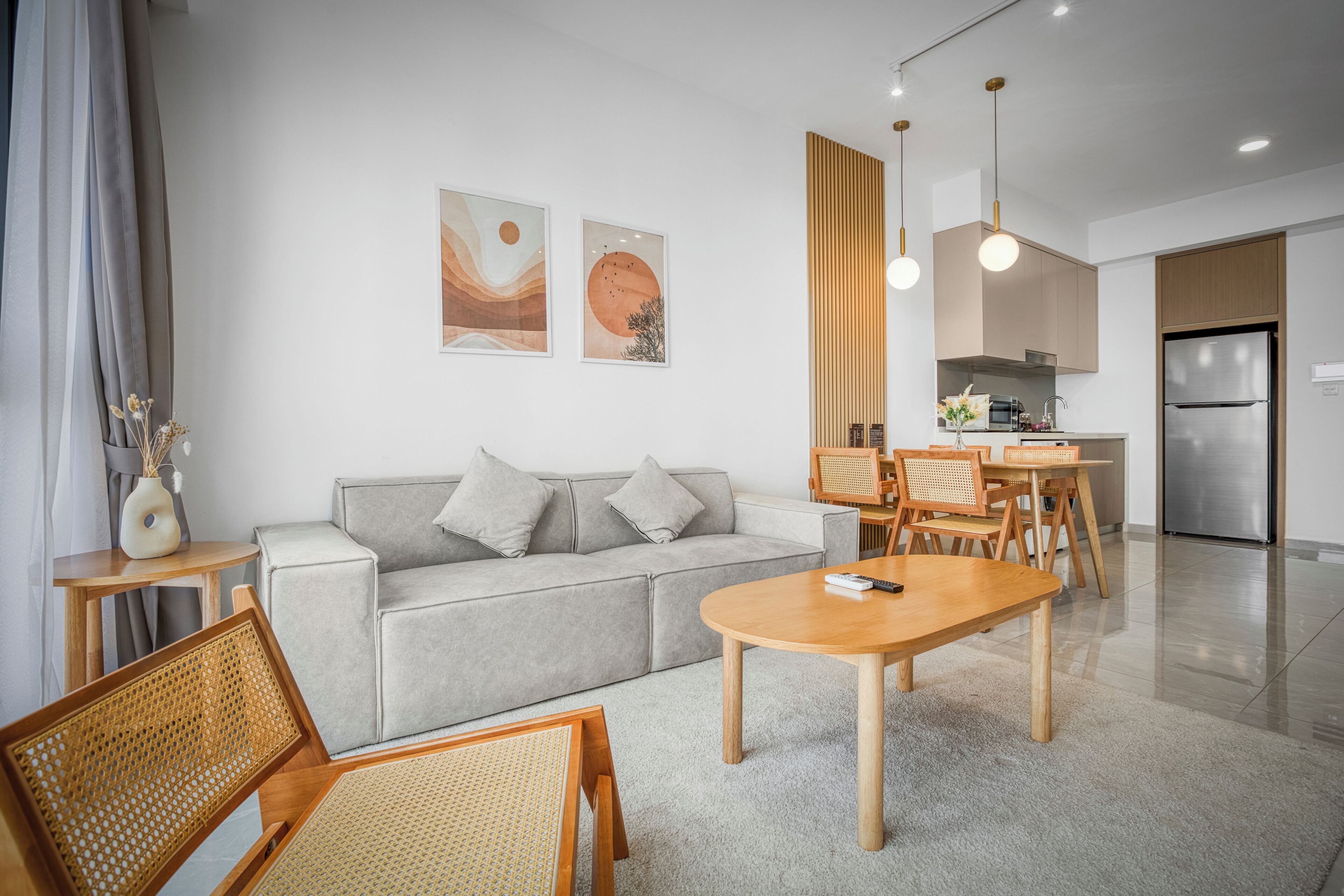 A modern living room with a grey sofa, wooden dining table, and chairs. There is a kitchen area with a refrigerator and wooden cabinets.