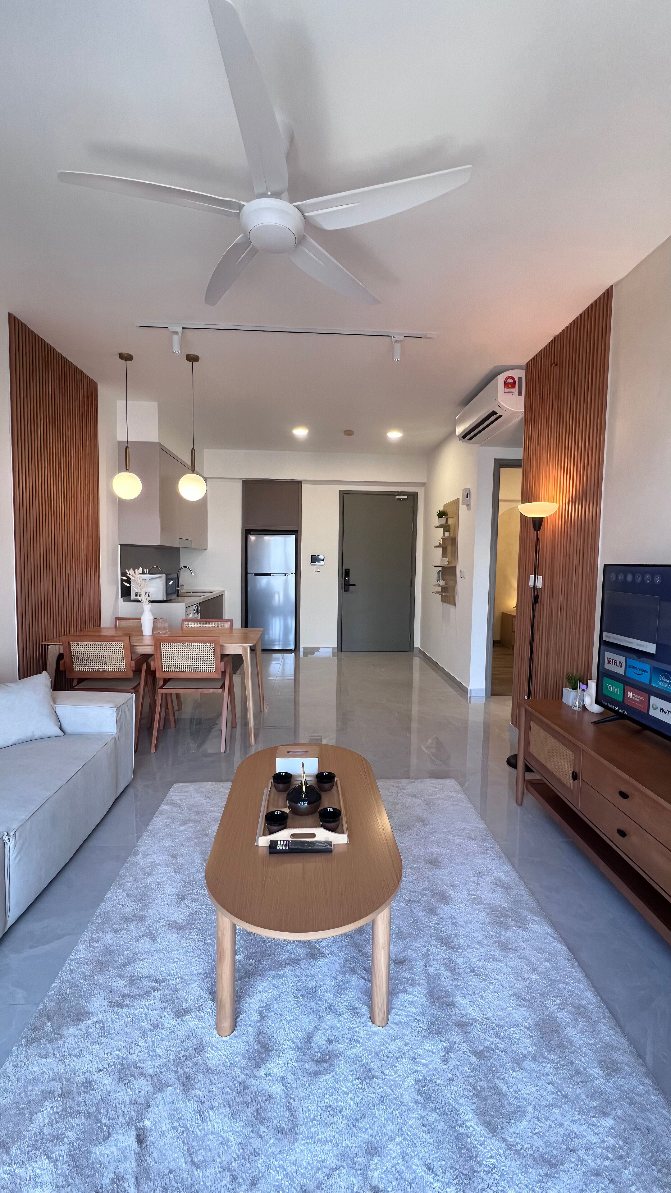 A modern living room with a wooden coffee table, a sofa, and a dining area with chairs. There is a ceiling fan, recessed lighting, and a flat-screen TV on a wooden cabinet.