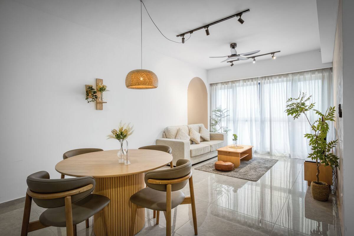 A modern living room with a round wooden dining table, comfortable seating, a ceiling fan, and large windows with sheer curtains.