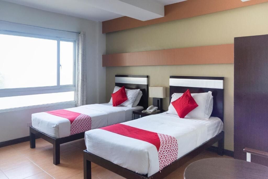 Een hotelkamer met twee bedden, elk met een rood-witte dekbedovertrek, een nachtkastje met een lamp en een raam dat natuurlijk licht binnenlaat.