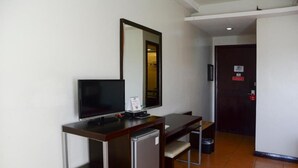 A compact hotel room with a single bed, a desk with a computer and a small refrigerator, a mirror, and a door with a red emergency exit sign. - EAST VIEW HOTEL (Bacolod)