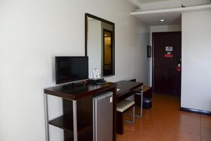 A compact hotel room with a single bed, a desk with a computer and a small refrigerator, a mirror, and a door with a red emergency exit sign.