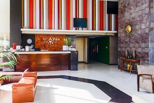 A hotel reception area with a wooden reception desk, a clock, and a television above it. There are two sofas and a small table with decorative items.