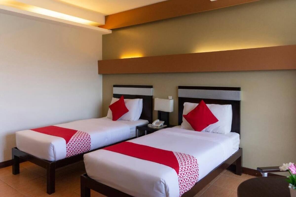 A hotel room with two single beds, each with a red and white patterned bedspread, a red pillow, and a bedside table with a lamp.