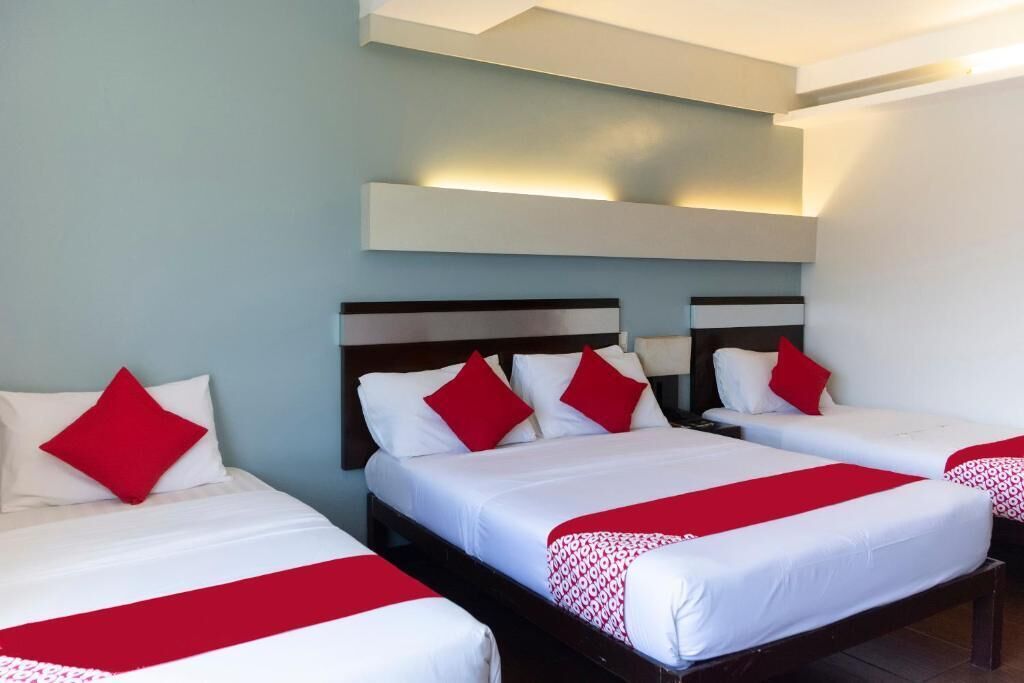 Quarto de hotel com três camas simples, cada uma com roupa de cama branca e detalhes vermelhos.