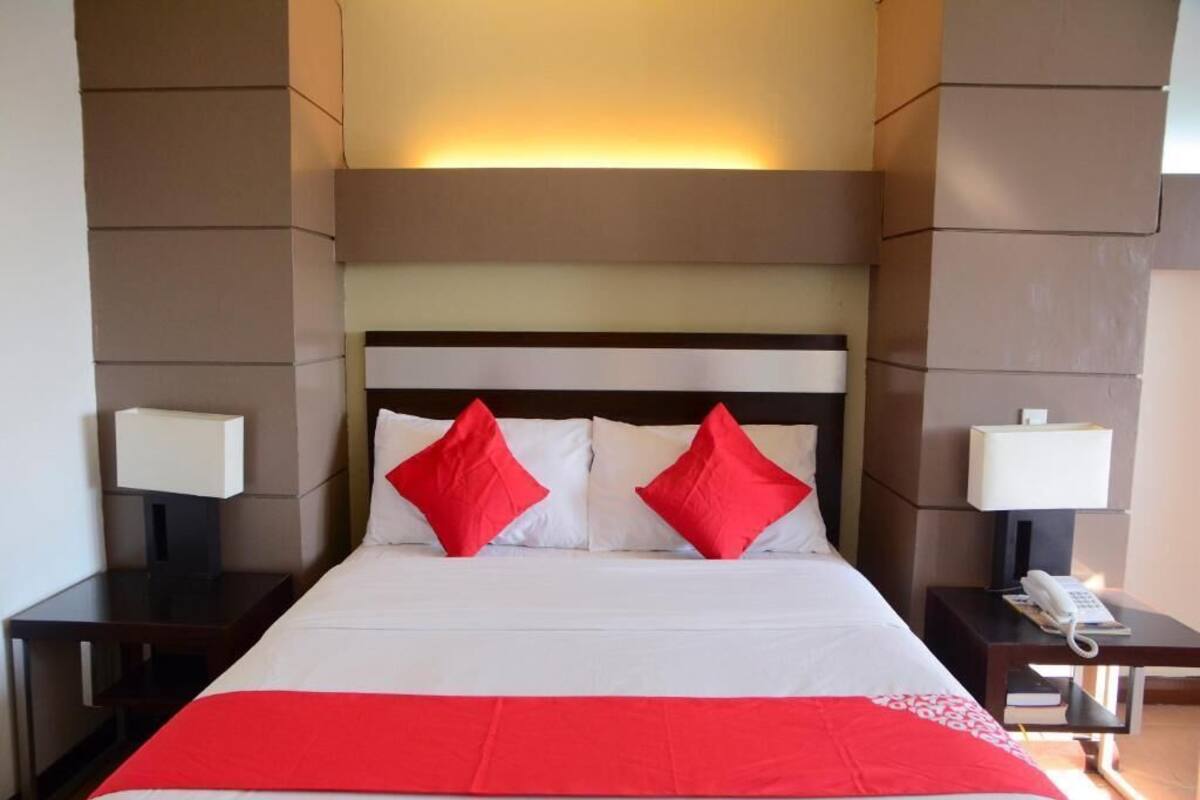 A hotel room with a large bed, two red pillows, a white bedspread, and a red blanket. There are two bedside tables with lamps and a phone on one of them.