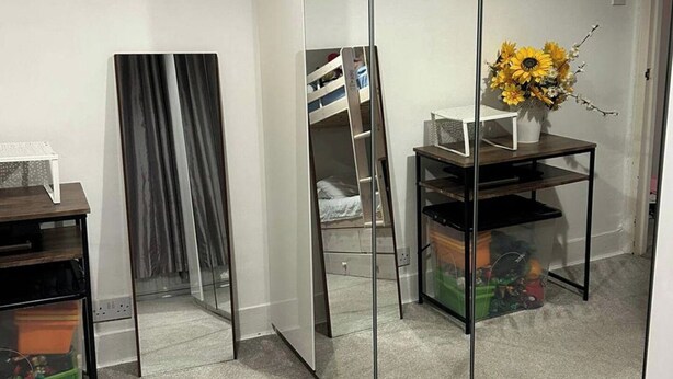 Une chambre avec une armoire à portes miroir, une petite table sur laquelle est posé un vase de fleurs et un lit avec une tête de lit blanche.