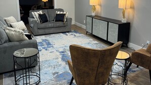 A modern living room with a grey sofa, a coffee table, and two armchairs. There is a console table with lamps and a rug with a blue and white pattern. - Amazing Home Away From Home (Valdosta)