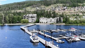 A marina with several boats docked, a bridge leading to a building complex, and a forested hillside in the background. - Top Floor Penthouse with Pool, gym and Boat Slip (Sicamous)