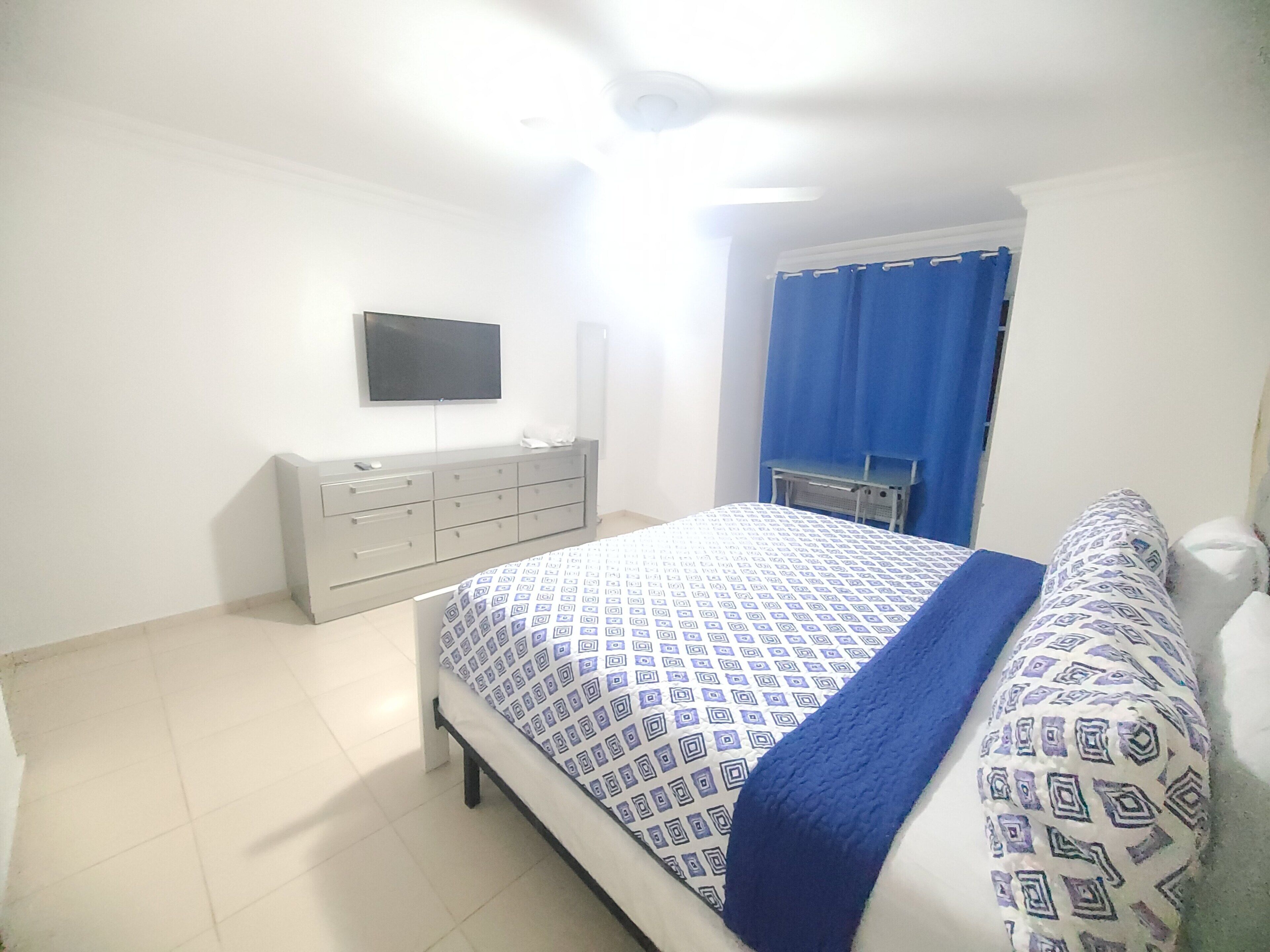 Un dormitorio con cama, cómoda, televisor en la pared y ventana con cortinas azules.