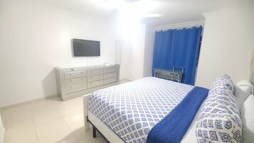 Un dormitorio con cama, cómoda, televisor en la pared y ventana con cortinas azules.